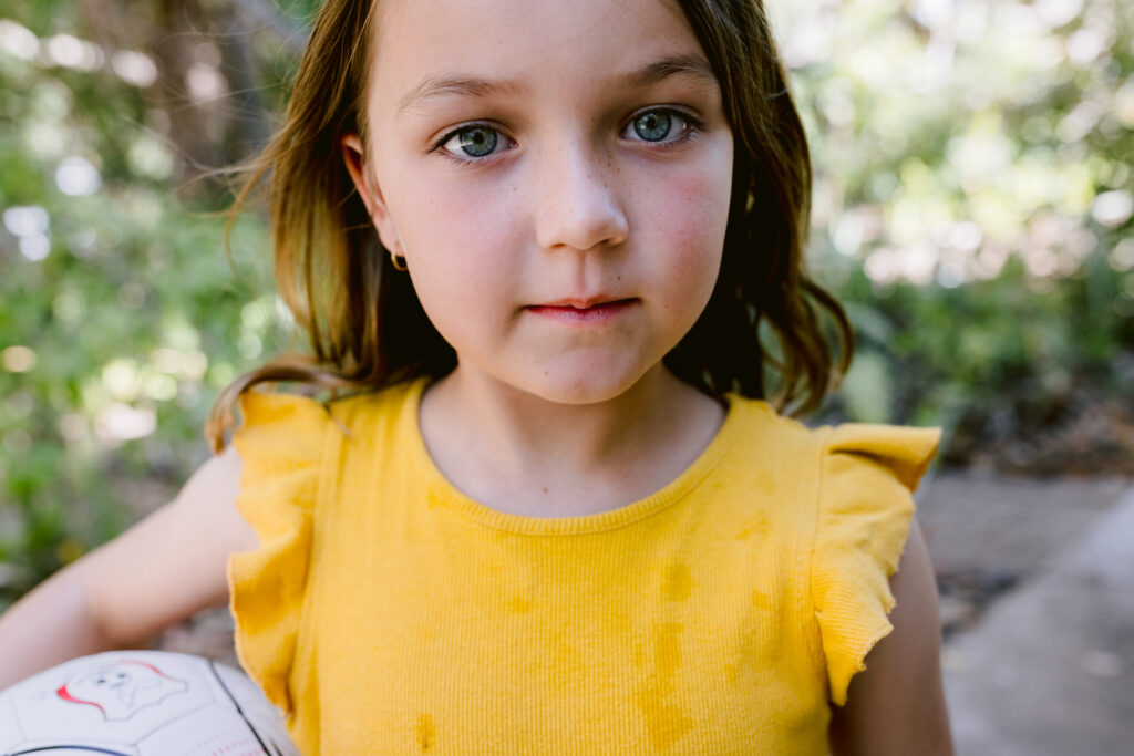 Photo of a young girl looking directly at the camera with a basketball tucked under her arm.