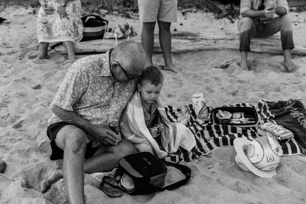 Grandfather sits next to his grandson on the beach towel and chats to him.