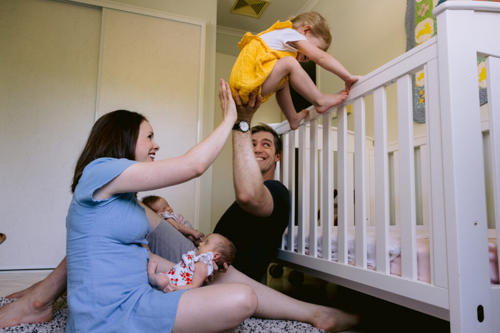 Mum and Dad each have a twin baby in their lap and with one hand each the help the toddler climb over the cot rail.