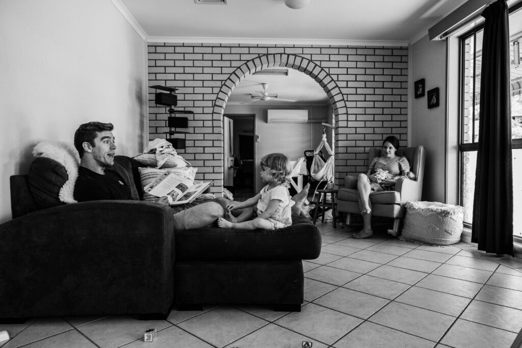 Dad reads a book on the couch to the toddler while Mum sits in the rocking chair with the twin girls.