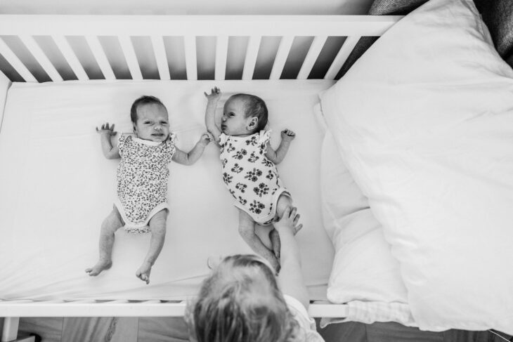 The big sister leans over the cot to touch the knee of her newborn twin sister