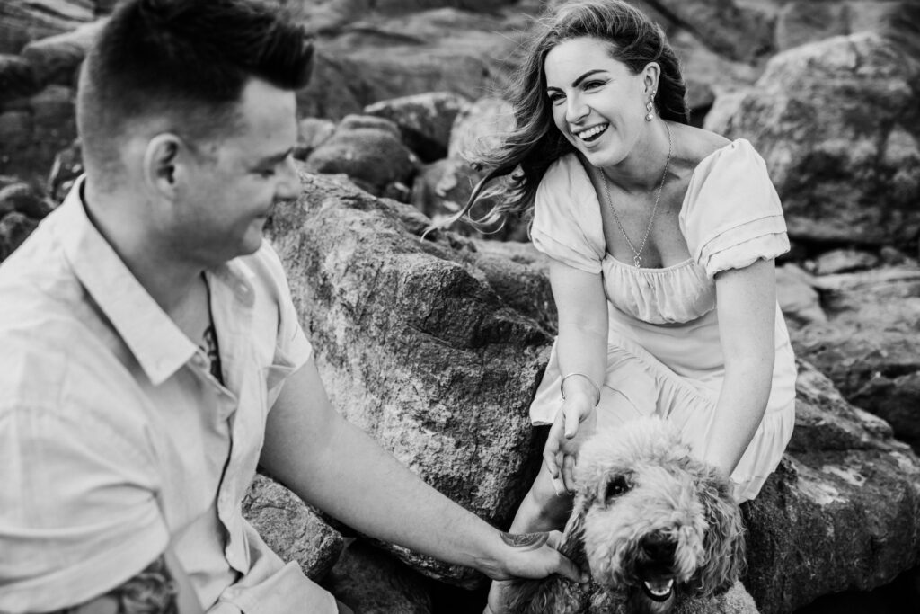 Trudy is looking at her fiancé Beau laughing as they sit on the rocks at the beach.