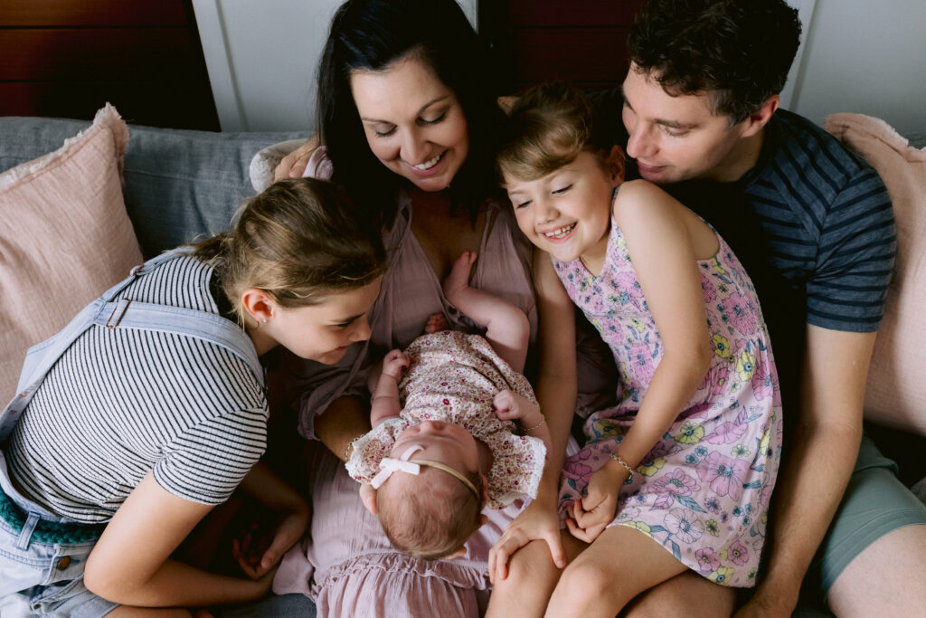 All the family snuggle on their couch at home looking at their newborn baby Verity.