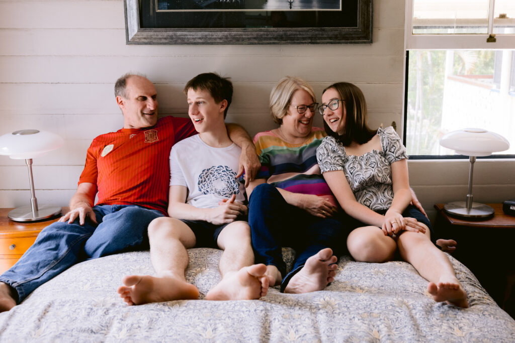 Father, Mother, Son and Daughter all sit on their parents bed laughing together.
