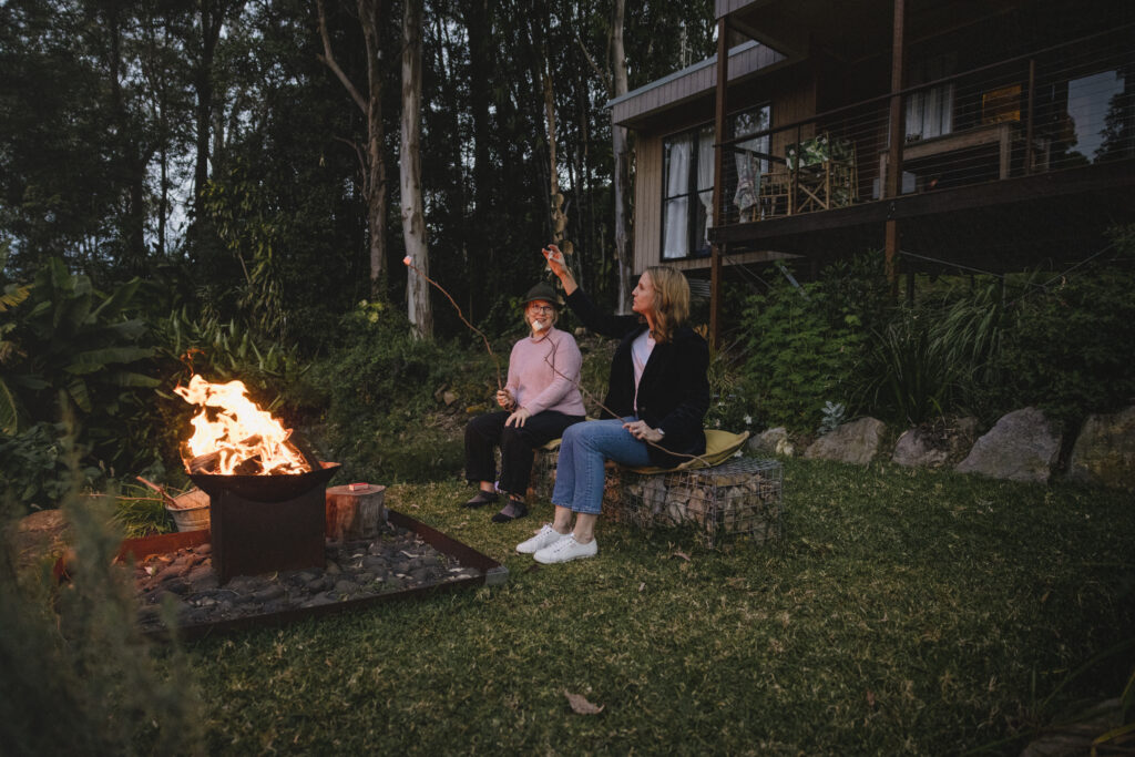 Katie and Elyse sit by the fire out the front of the Airbnb The Wilds Container Home roasting marshmallows.