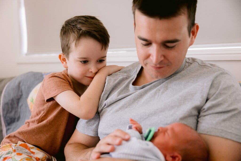 Big brother leans on his Dad's shoulder looking lovingly at his newborn baby brother.