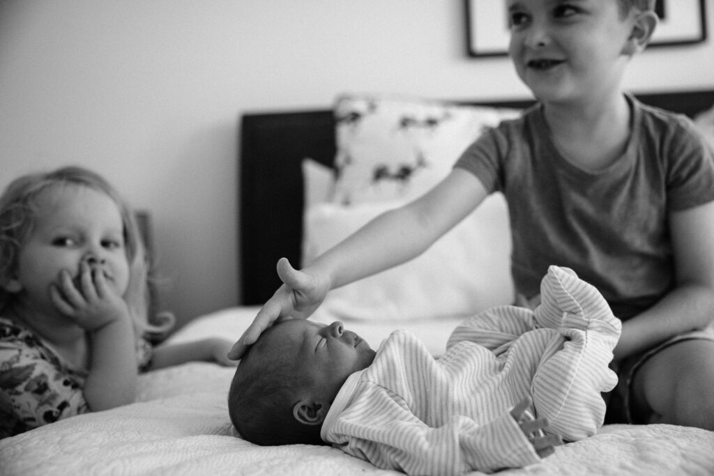 Newborn baby lying on the bed with big brother gently touching his head and big sister leaning on the bed watching on