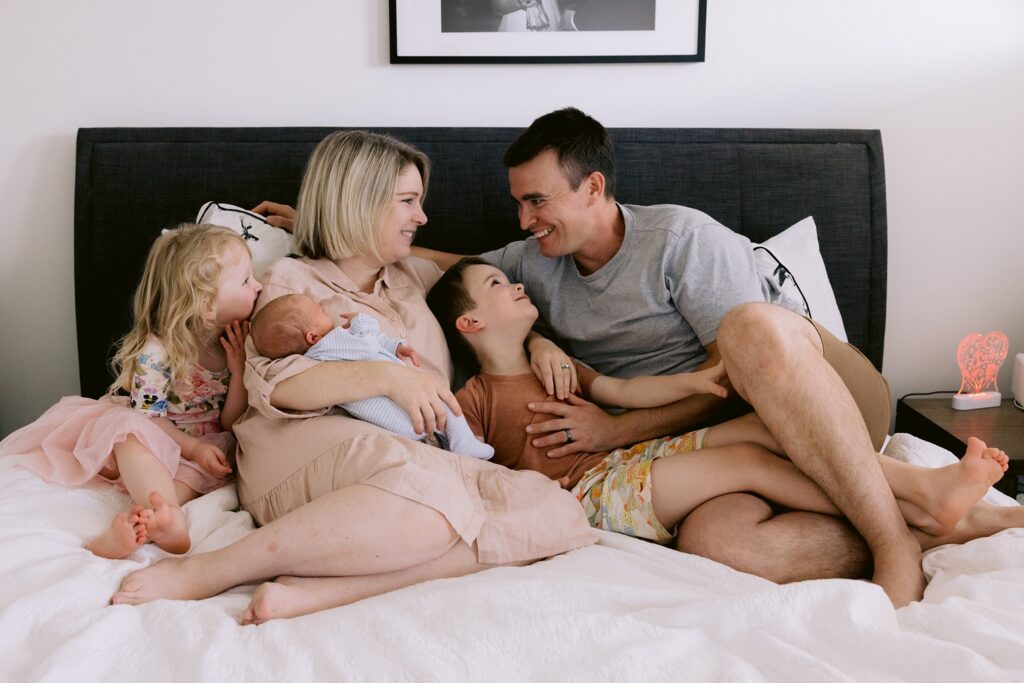 Family photo of Mum, Dad, Big brother, Big sister and newborn baby all snuggled on the bed at home.