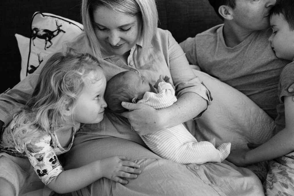 Mum is holding the newborn and the sister give a cheeky look as she snuggles her newborn baby brother. The son cuddles into his Dad's shoulder.