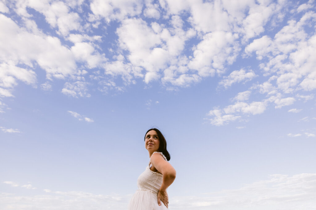 Maternity photo of a pregnant lady waiting a white dress looking back over her shoulder with a blue sky filled with clouds behind her.