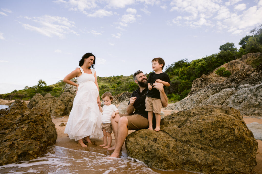 Maternity Family photo. Mum is standing holding the youngest childs hand, while Dad is sitting on the rock with his feet in the ocean holding onto their other son who is standing beside him. 