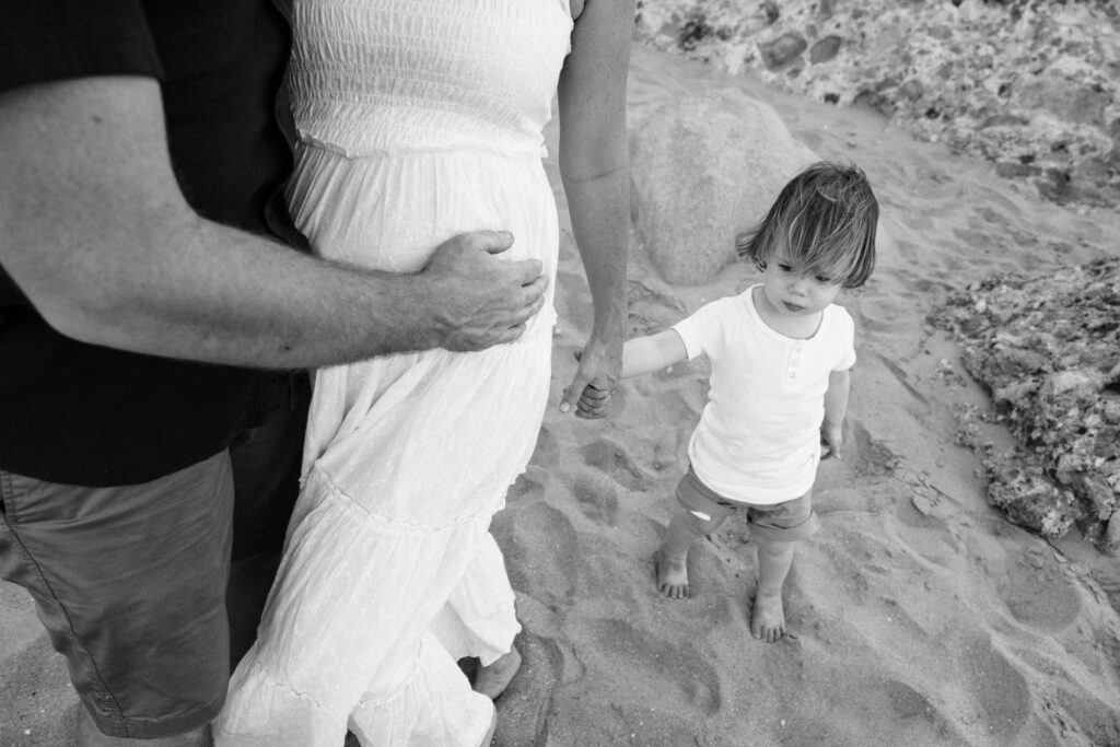 Dad puts his hand on the mum's pregnant belly, while she holds her youngest childs hand.