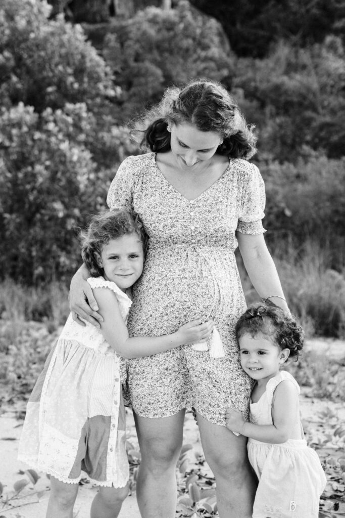 Maternity photo of Mum with her two daughters cuddling her. They are standing on the beach with bushes behind them.