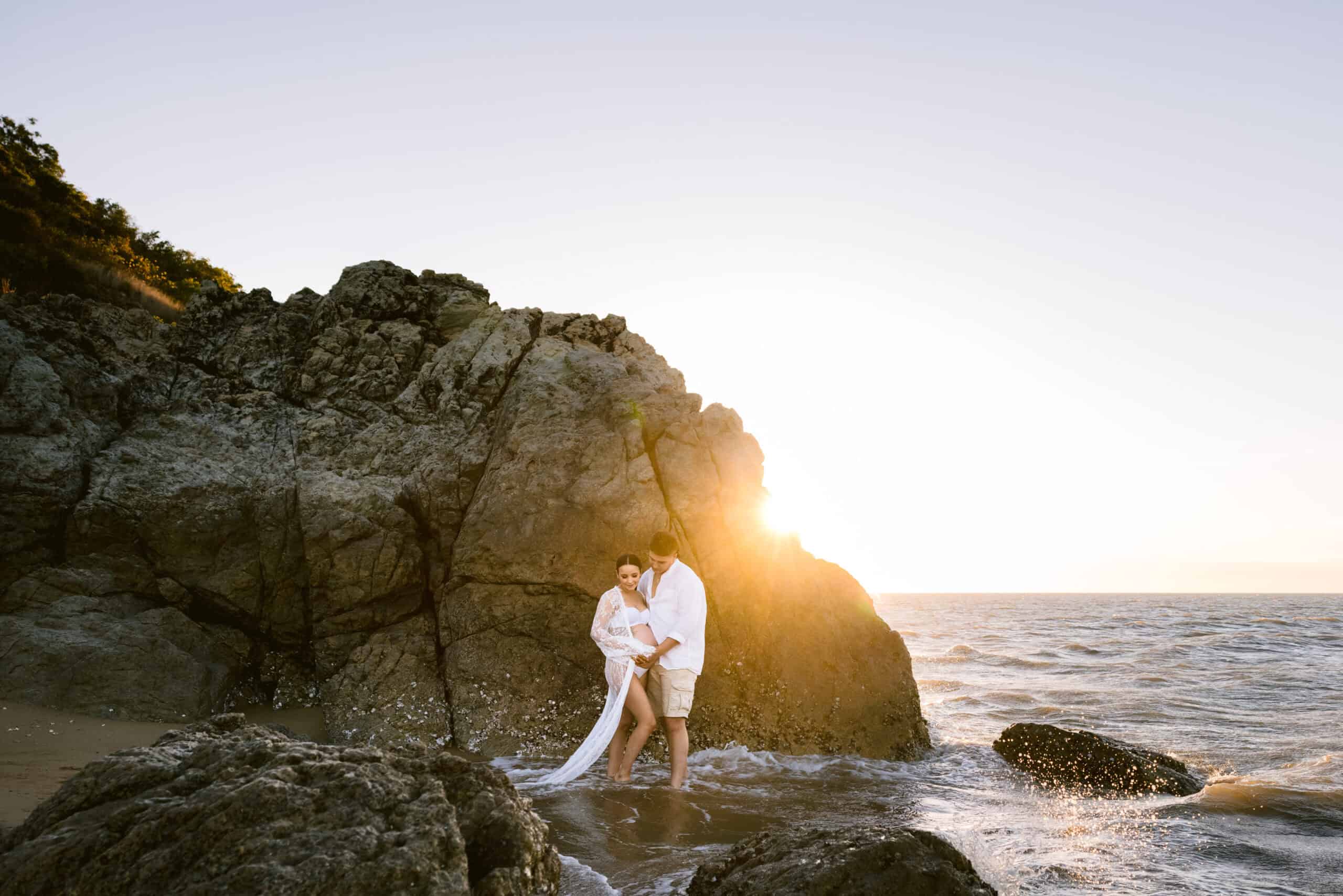 A maternity photo of a pregnant lady and her partner on the beach with a large boulder behind them with the sun shining over the corner of the rock.