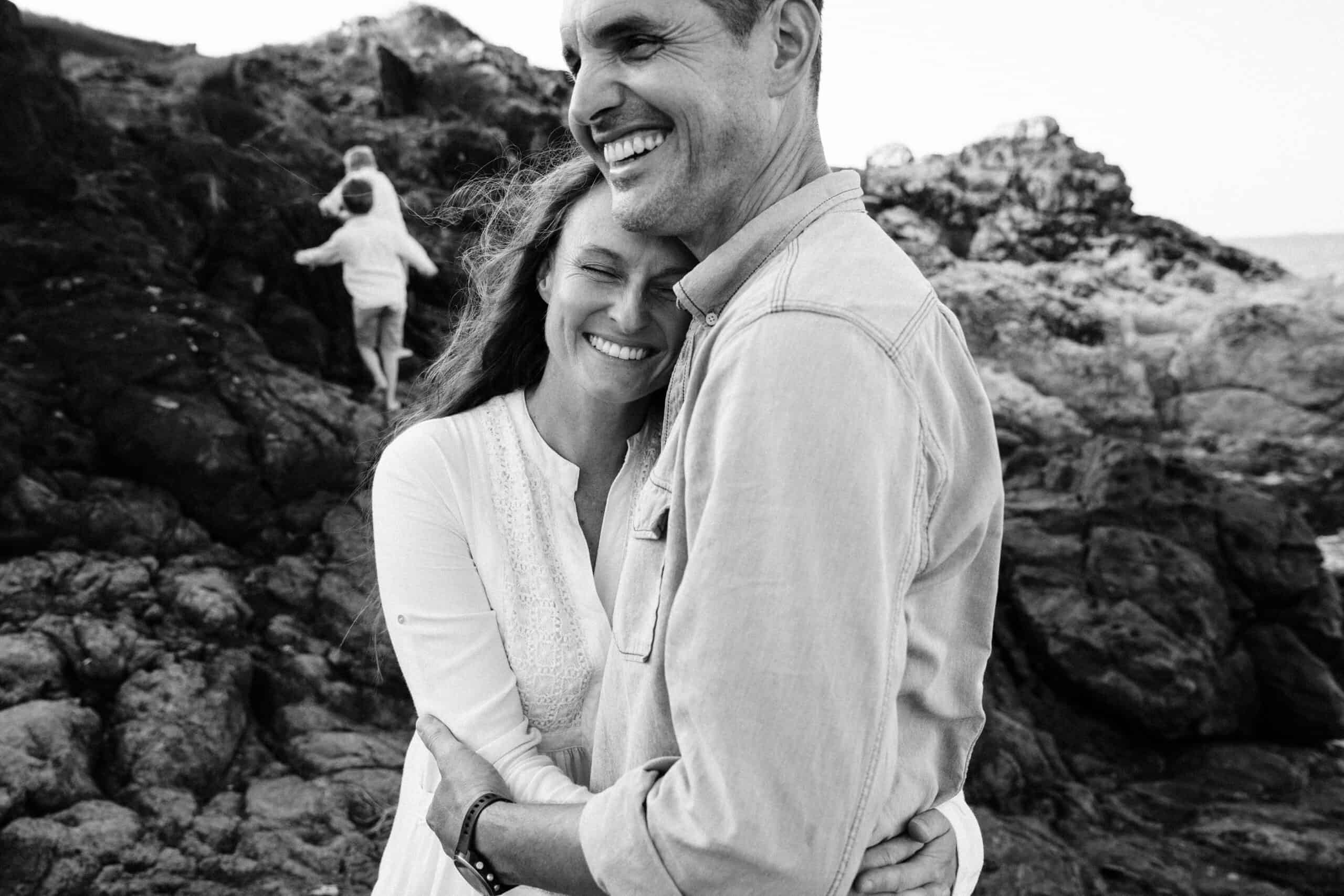 A husband and wife stand with arms wrapped around each other with faces that beam happiness while their sons climb on the rocks in the background.
