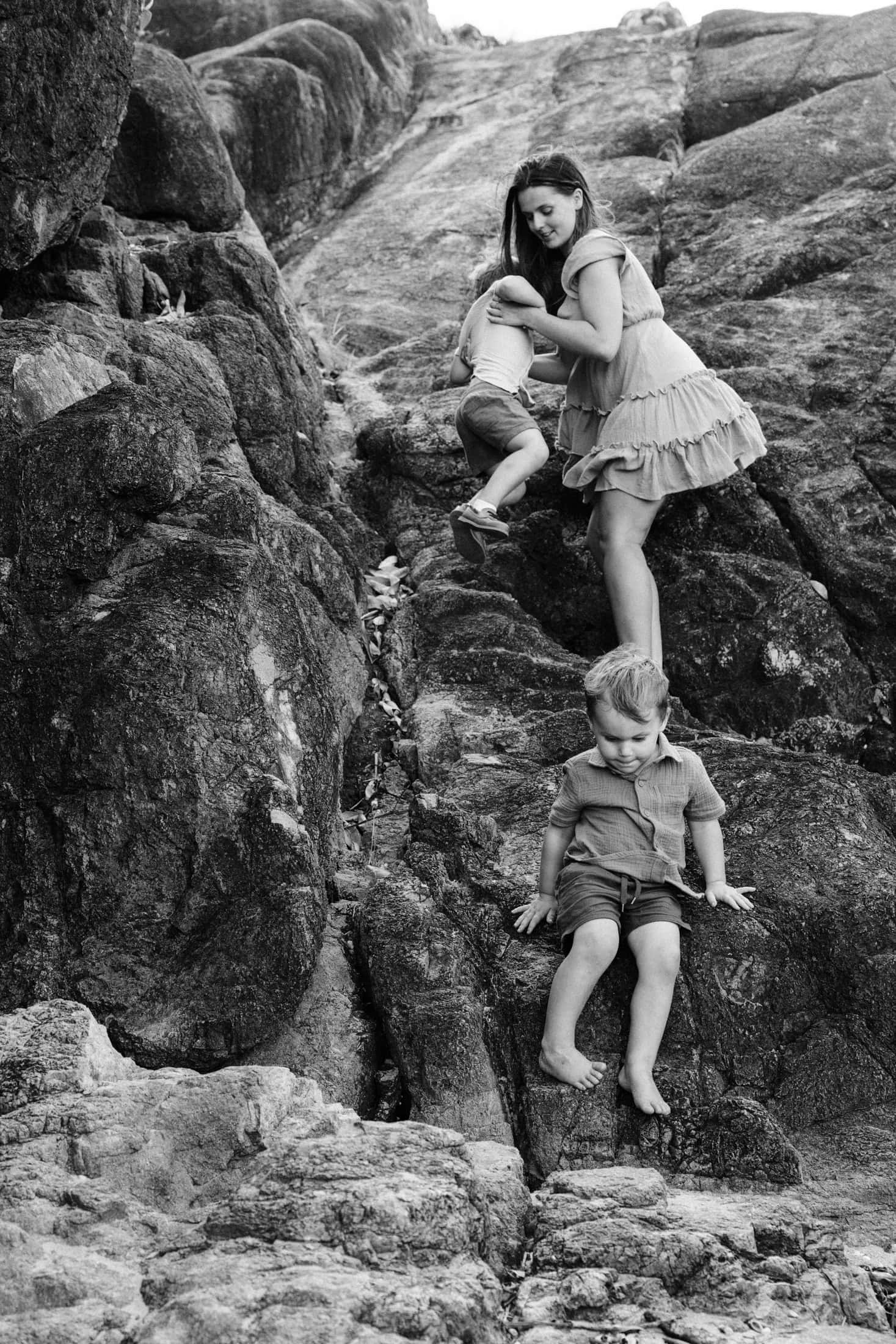A mother helps her two little boys whist they climb the big rocks by the beach.