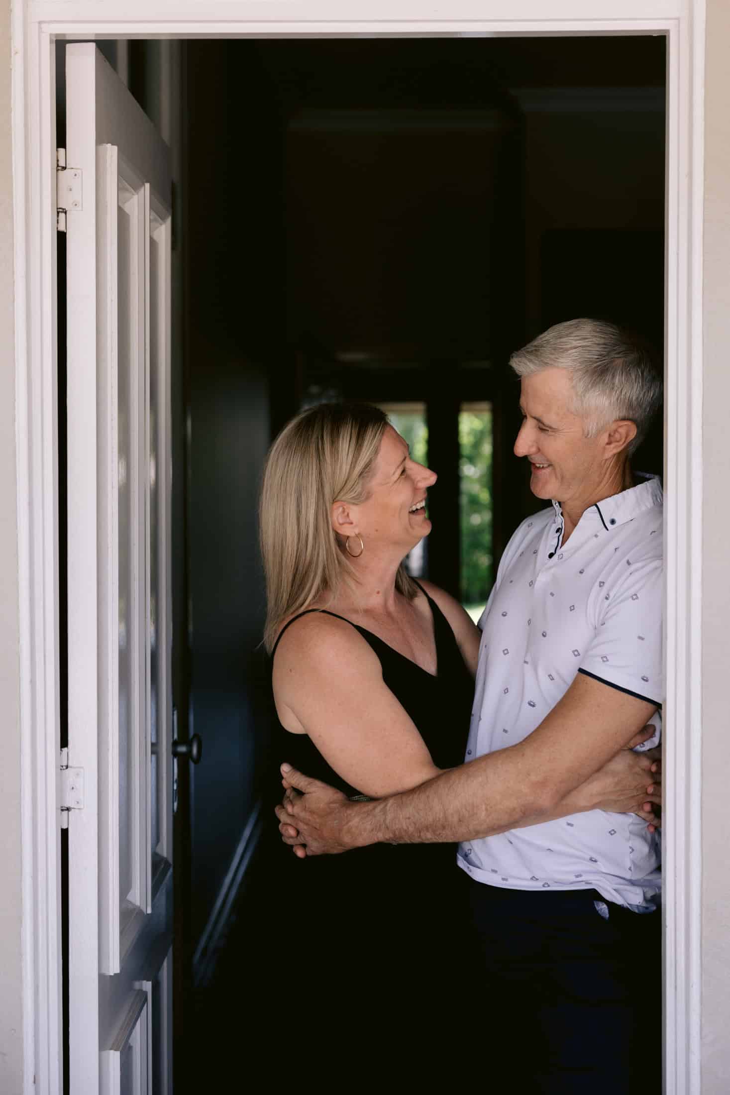 A husband and wife stand with their arms around each other looking towards each other in the doorway of their old Queenslander home in Townsville.