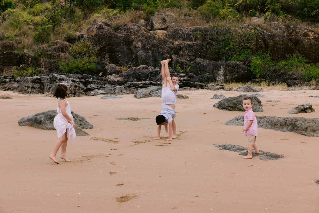 Four children play at the beach. One brother holds another legs in the air as he handstands.