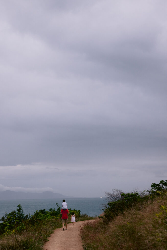 A Dad carries his child with a sore foot on his shoulders along a dirt path by the beach. The sky is getting dark as the sun sets.