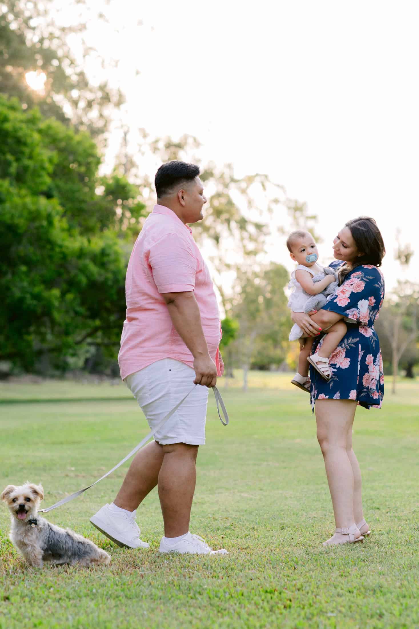 Mum hold their young daughter and Dad is walking over to them in the park with their dog