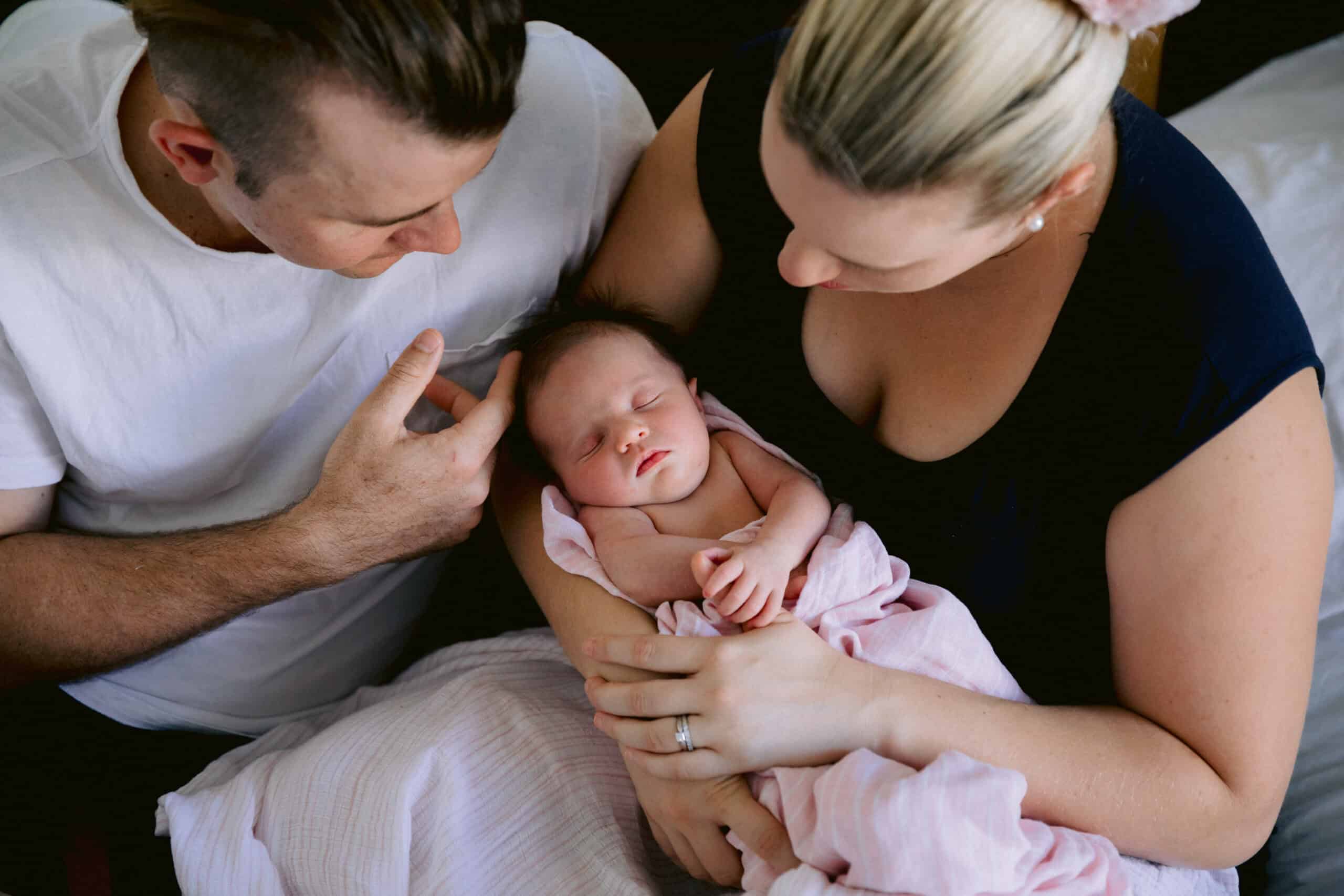 A newborn is being held by her mother. Both parents are looking at their new baby girl and Dad is gently touching her hair.