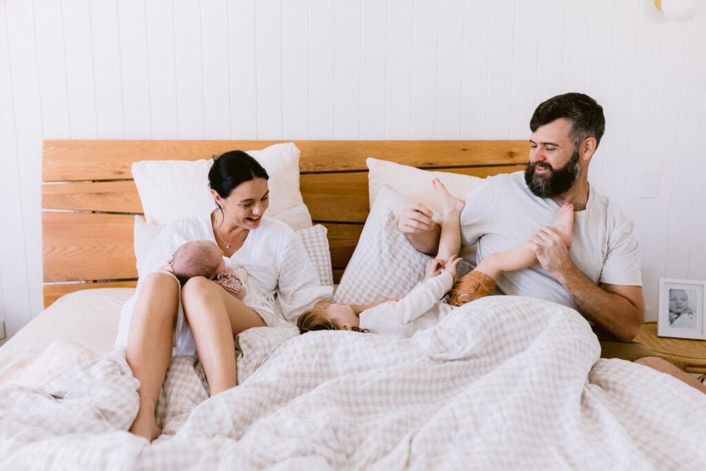 Mum is holding the baby girl and Dad is wrestling with the brother on the bed