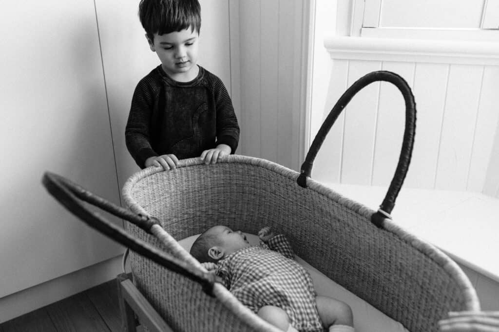 The big brother is looking at his baby sister in the basket.
