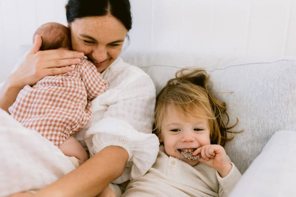 Mum is holding the new baby and her young son is snuggled next to her eating a chocolate freckle.