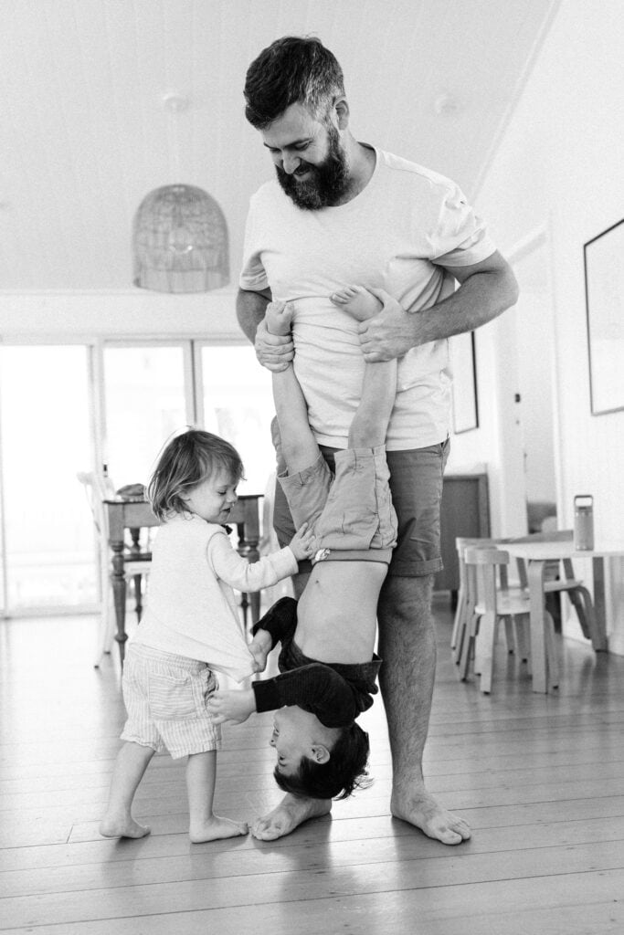 Dad is playfully hanging one son upside down while the younger brother joins in on the fun
