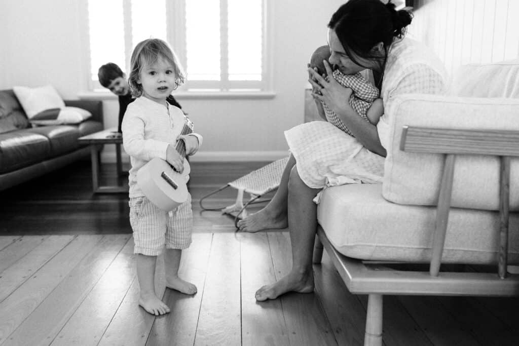 Mum is holding the baby and her young son is holding a guitar in their lounge room