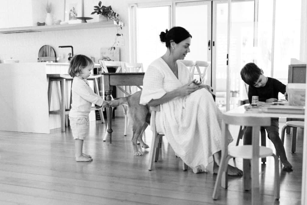 Mum is sitting at a little table playing with her son. The other brother is gentle holding the dogs tail.