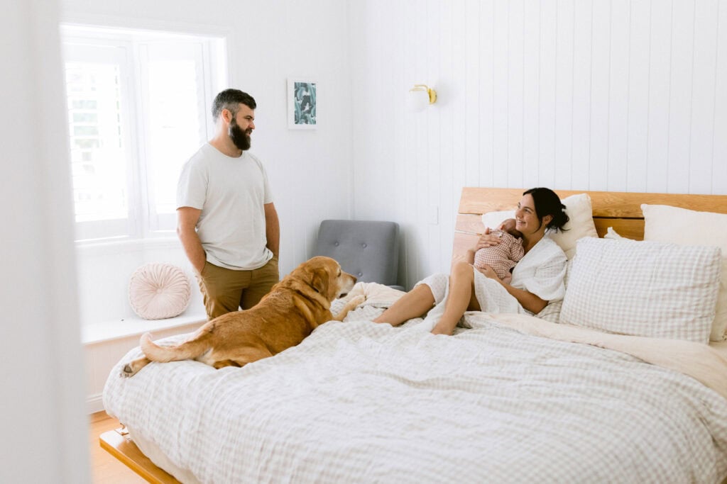 Mum is sitting on the bed holding the newborn baby girl . Dad is standing beside the bed talking and smiling. Their dog is lying on the bed.