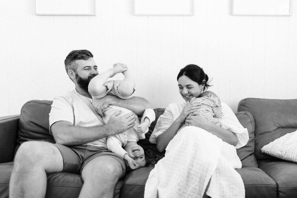 Dad is wrestling his son on the couch at home, while mum looks on smiling siting next to them and holding the newborn baby girl