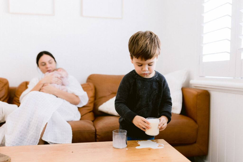 Mum is holding baby on the couch and the big brother is drinking his milk which is dripping off his lips and has spilt on the coffee table 
