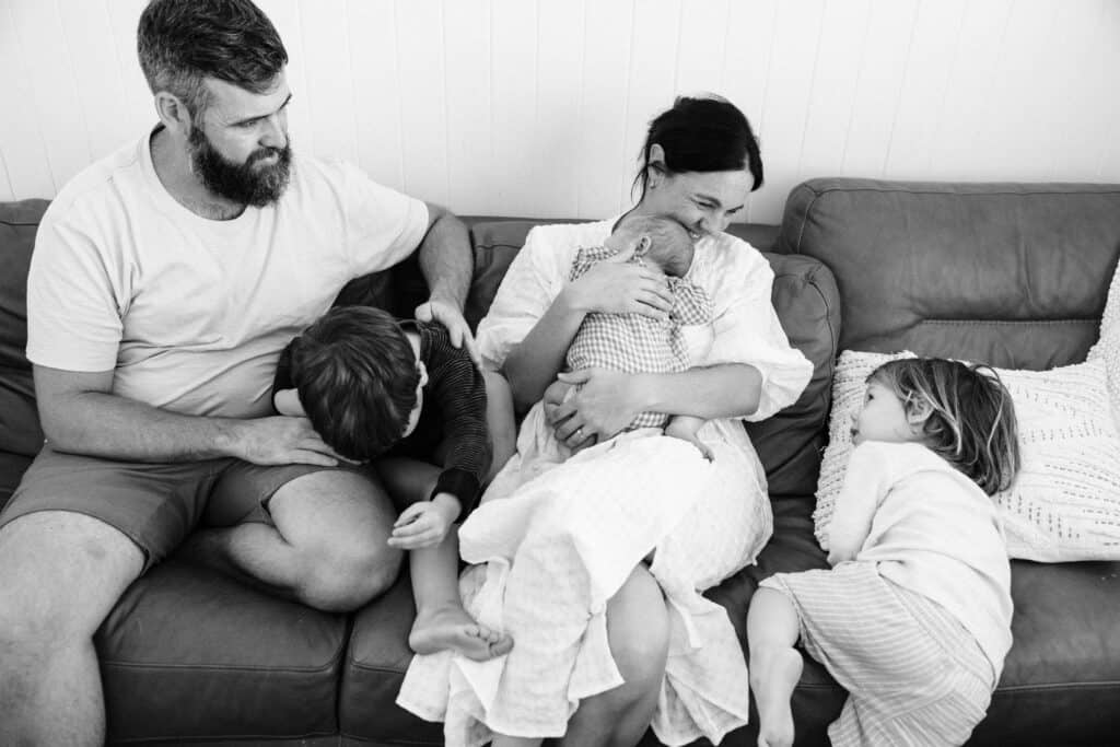 A family is sitting on the couch. Mum is holding the baby and Dad is wrestling one brother while the other brother is climbing ono the couch. Mum is looking at her son smiling.