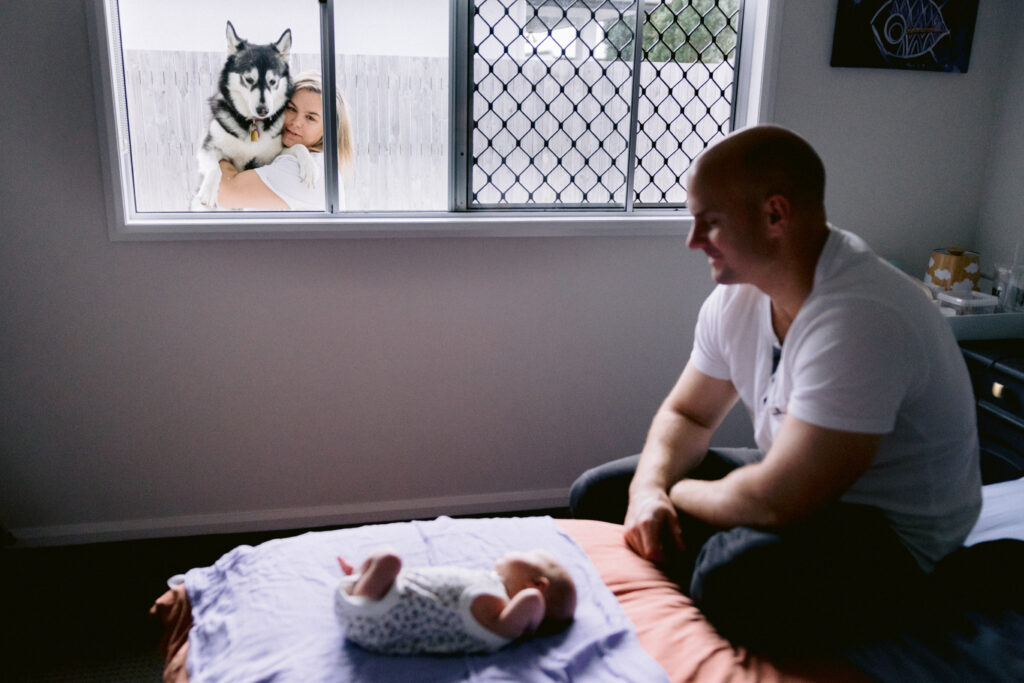 The mother holds up one of the dogs to look through the window at the Dad and baby