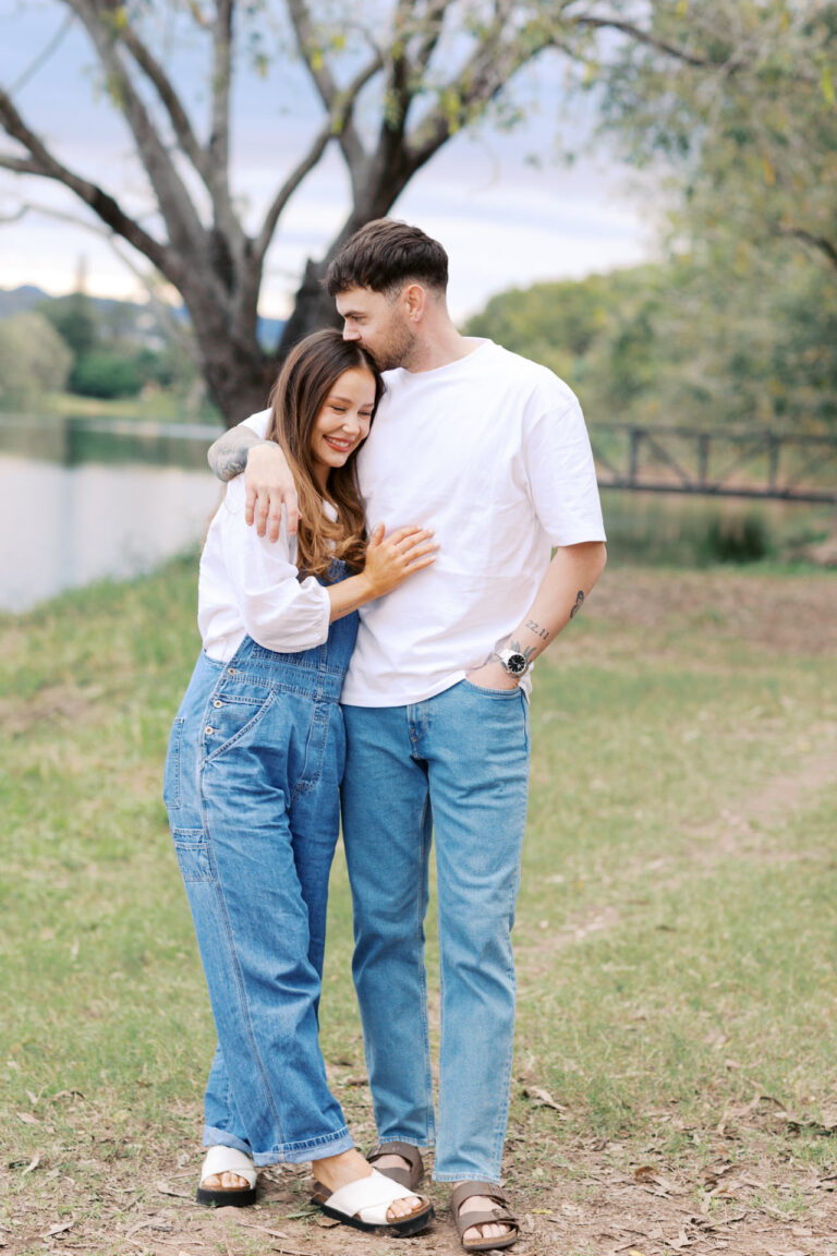 A couple stand next to each other by the river as she snuggles into him and he kisses her head