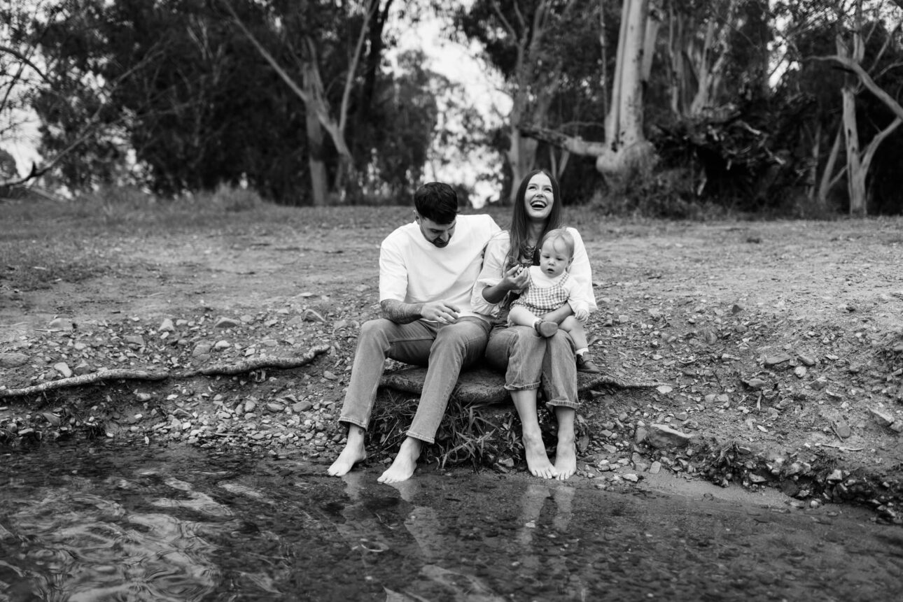 a Mother and father sit on the bank of the river with their little boy laughing