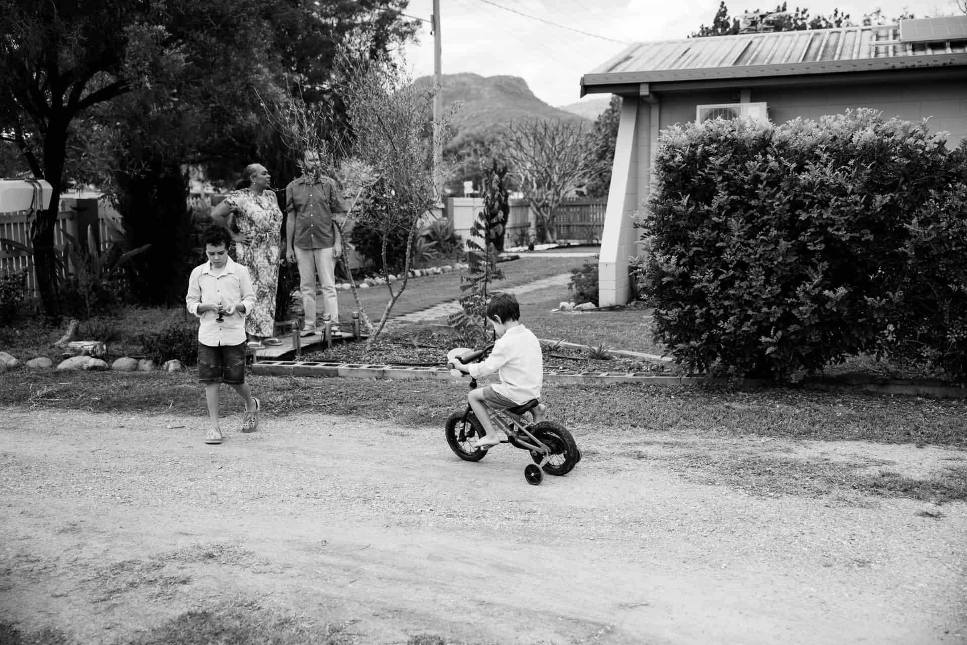 Black and white photo of a family in their yard. Mum and Dad are looking a tree, one son is looking at something in his hands and the other young son is riding his bike with training wheels on