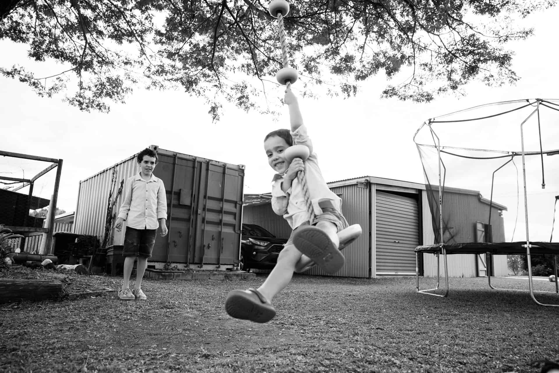Two brothers are under a big tree in their yard. Playing on a swing.