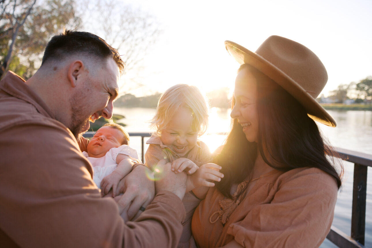 A family photo of a husband and wife holding their newborn daughter and three year old son. The glow of sunrise is shining through