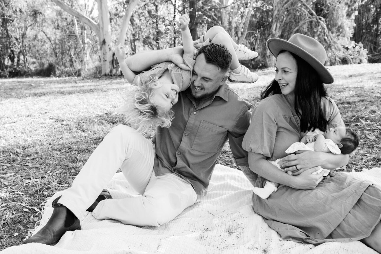 A father is playfully swinging his son over his shoulder whilst sitting on a blanket on the grass with his wife who is holding their newborn
