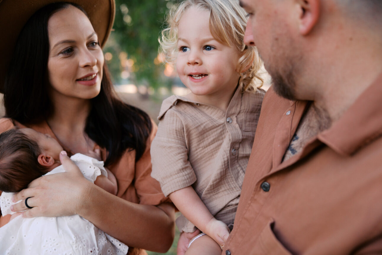 Mum is smiling holding her newborn while looking at her three year old son who the father is holding