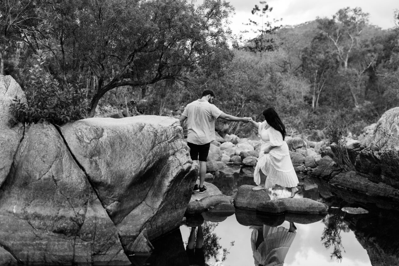 A maternity photo shoot, of a husband and wife. The husband is holding his wife's hand as they walk across rocks in the creek