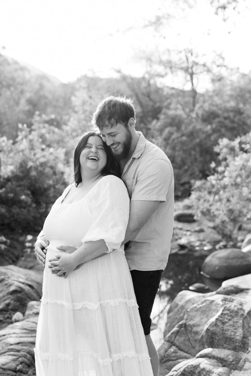 A husband stands behind his pregnant wife with his arms wrapped around her and they are both laughing