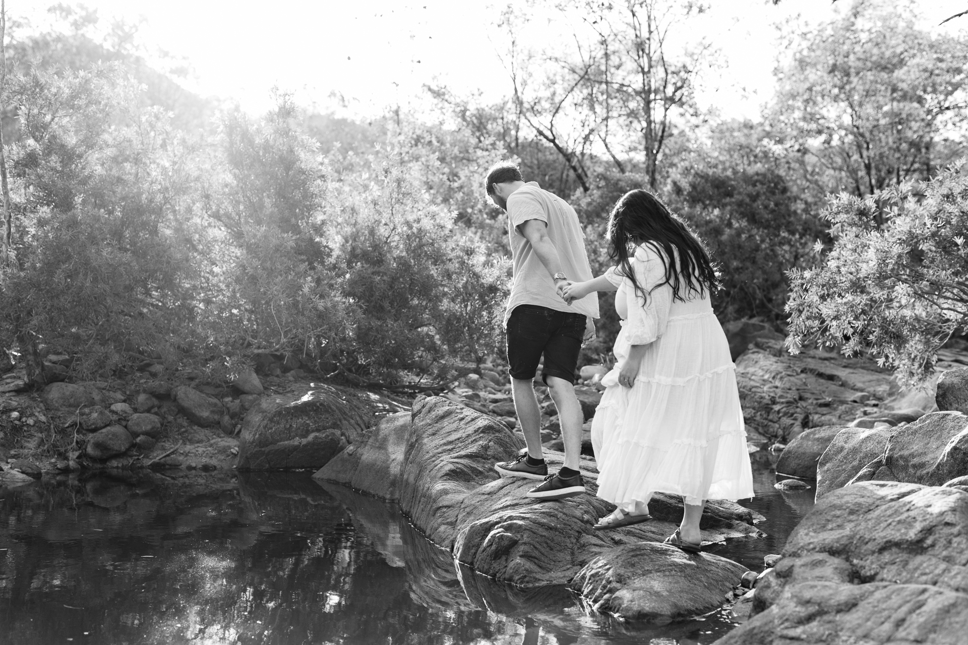 a husband is holding his pregnant wife's hand as they walk over a rocky creek bed